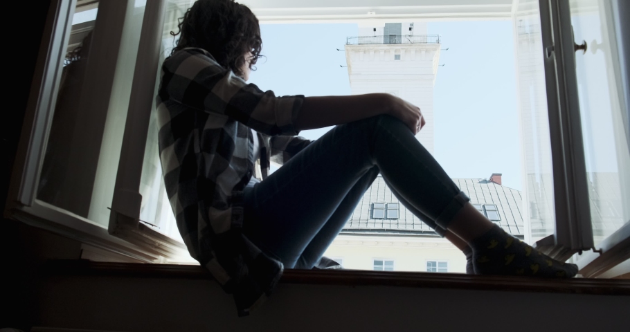 Young woman sitting on window sill and enjoying old town view. Silhouette of relaxing Girl on fresh air at home