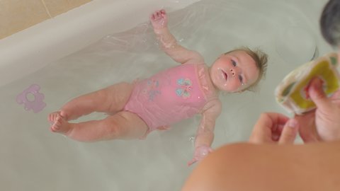 Infant baby floats independently in bathtub. Swimming lesson for baby at home. Parent is watching, top view. Swimming skills training from an early age.
