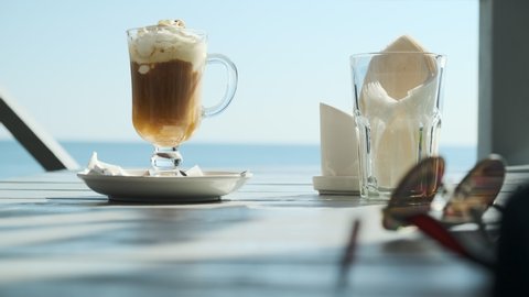 beautiful view of glass with iced coffee with ice cream on background of magnificent view of ocean or sea, concept of beach holiday or freedom, next to it there is glass with napkins and sun glasses.