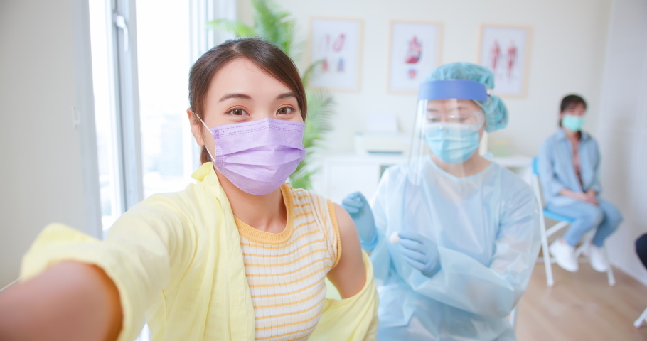 Smiling Asian woman wearing face mask taking selfie with mobile phone while receiving coronavirus vaccine Royalty-Free Stock Footage #1077297359
