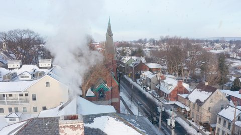 Snow falls. Cold snowy morning. Aerial establishing shot of small town community. Car passes by on street as smoke billow from chimney.