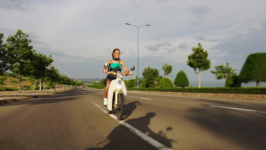 Carefree Caucasian Fashionable Model Sticking Tongue Out and Smiling While Riding White Moped Scooter On Open Road During Solo Roadtrip, Slow Motion