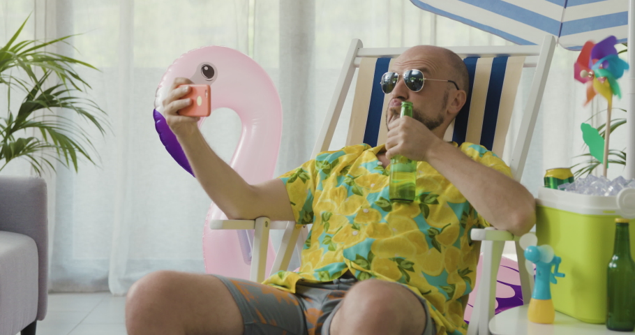 Happy man spending his summer vacations at home alone, he is sitting on a deck chair in the living room and taking selfies with his smartphone Royalty-Free Stock Footage #1077309725