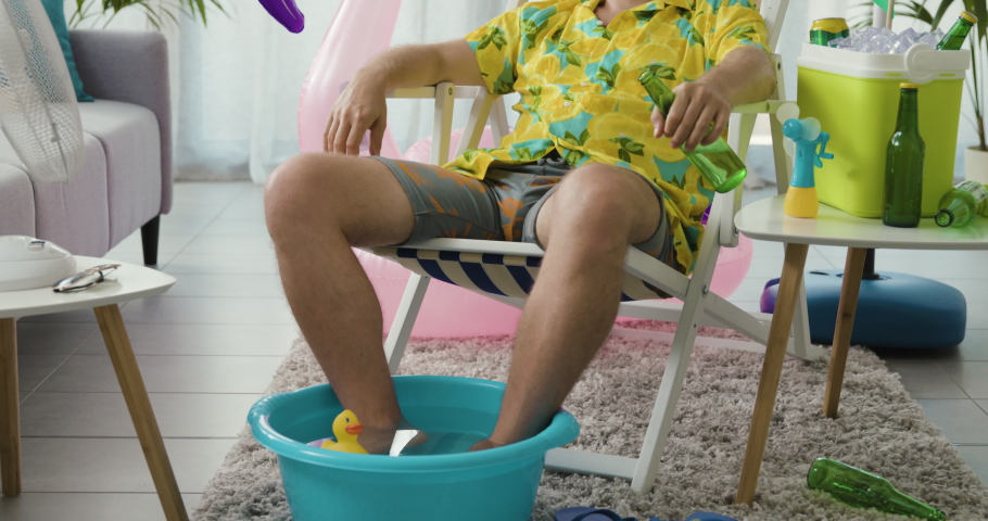 Man staying at home for the summer and suffering from heat, he is sitting on a deckchair in the living room and holding a beer Royalty-Free Stock Footage #1077309737