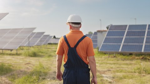 Maintenance assistance technical worker in uniform is checking an operation and efficiency performance of photovoltaic solar panels. Construction engineer walks between solar panels on field station.