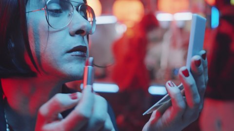 Portrait of transgender man in brunette wig applying red lipstick and looking at camera while sitting in bar with neon light