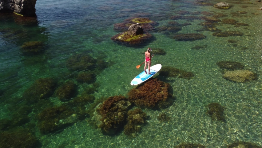 Aerial drone view on young attractive brunette woman with long hair in red swimsuit, swimming on sup in calm sea around volcanic rocks, like in Iceland. Summer holiday vacation and travel concept.