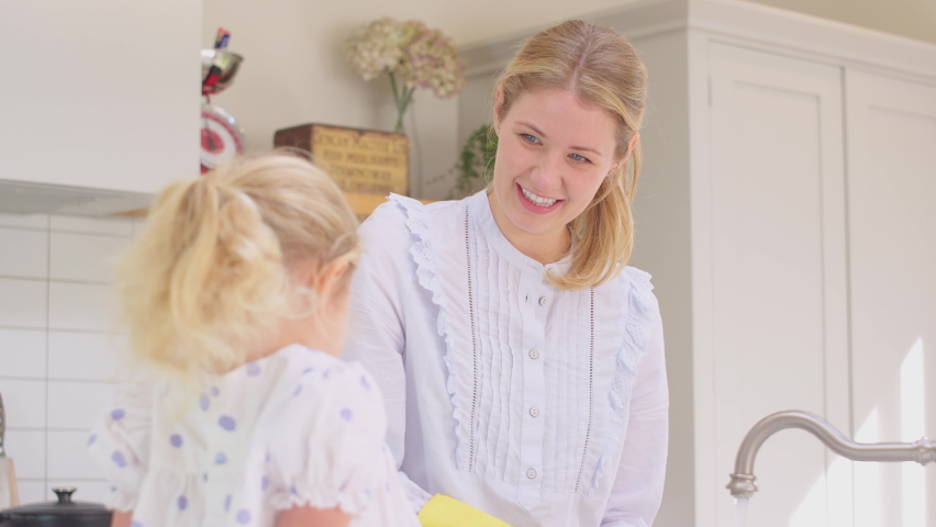Mother wearing rubber gloves at home in kitchen with young daughter having fun and washing girl