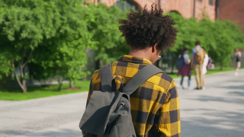 Follow shot of afro-american male student reading book walking outdoors and looking back at camera. Happy african guy with book smiling at camera outside college building