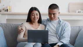 Cheerful Korean Couple Using Laptop Computer Together, Nodding Head In Approvement Sitting On Couch At Home. Japanese Family Browsing Internet. Great Website Concept. Zoom In