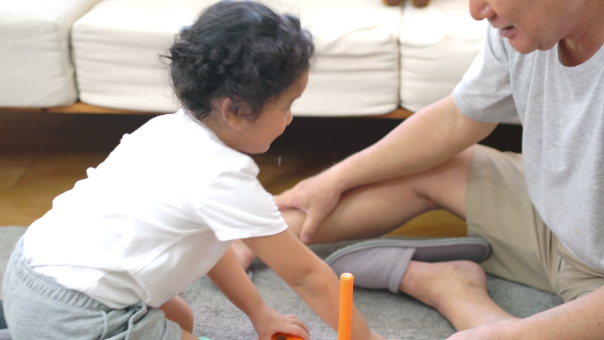 Happy Asian family having fun together at home. Smiling grandfather play toys with little grandchild girl in living room. Retired senior man teach cute baby granddaughter play colorful wooden block.