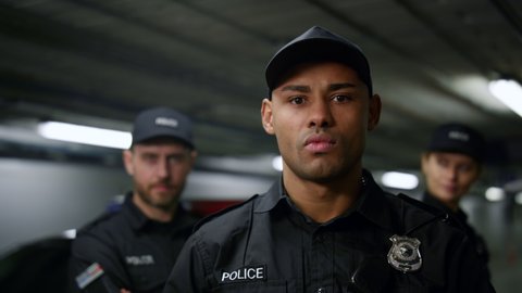 African policeman with serious face posing at camera. Brutal police officer in cap standing at patrol car with team on background. Portrait of male cop looking at camera 