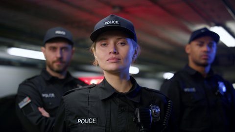 Portrait of serious policewoman in cap posing at camera with colleagues on background. Female cop standing at car with flashing lights. Police officer looking at camera 