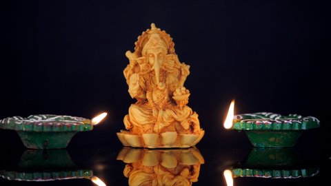 A handmade clay idol of a Hindu God Ganesha for the festival of Ganesh Chaturthi. Closeup shot of two colorful Diyas burning brightly on a glass table against a black background