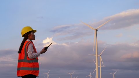 Young female engineer using walkie-talkie to checking system against wind turbine farm.