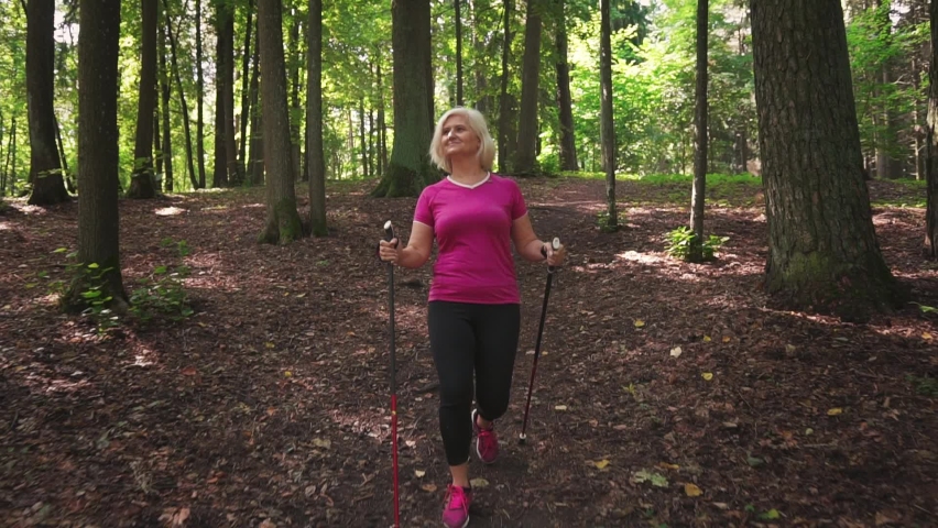 
Mature Caucasian woman doing Nordic walking in the forest. Active sports pensioner walks in the park with Nordic walking sticks, slow motion.	
