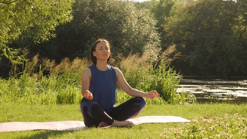 Young woman doing yoga exercises in summer city park. Health lifestyle concept. Portrait of peaceful female meditating in the morning, enjoying sunrise. Lady practicing yoga, sitting in lotus position