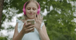 Happy teenager making online video call with modern pink headphones in park 