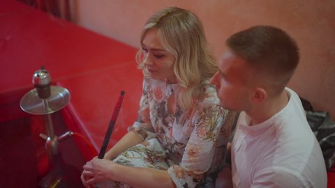 High angle view of relaxed beautiful slim young woman smoking hookah as man covering lady with blanket. Happy relaxed Caucasian couple resting in hookah bar indoors. Slow motion