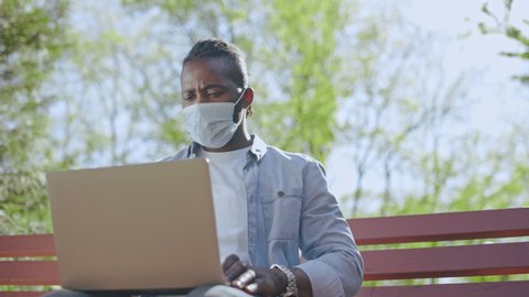 Young man in mask working on laptop while sitting in park, remote work