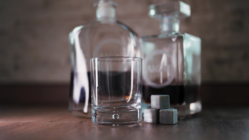 Barman pouring golden whiskey, cognac or brandy from bottle into glass on table. Bartender pouring glass of alcohol drink bourbon, rum and adding ice cooling stones.