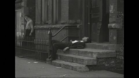 1940s: Man sleeps on stoop of building, children walk by, man appears from alley and stands watching. Boy approaches sleeping man on stoop, holding stick.