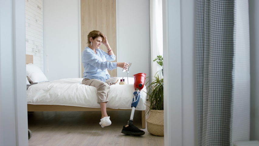 Disabled mature woman sitting on bed indoors at home, looking at camera. Morning routine with leg amputee concept.