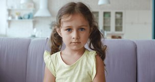 Adorable preschooler girl with two ponytails wearing yellow t-shirt sitting on the sofa in the living room , seriously posing looking at camera.. 4K video footage
