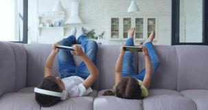 Cute kids, lying on a comfortable sofa with their legs raised, play with digital mobile phones. Schoolboy with headphones resting on the couch next to his younger sister. 4 K video footage