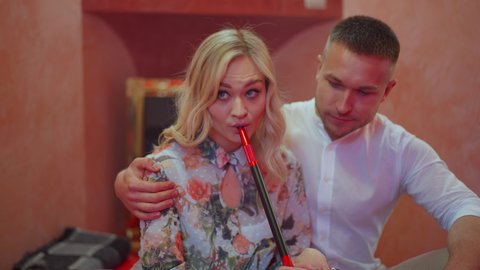 Portrait of young blond beautiful woman smoking hookah as man hugging and talking in slow motion. Relaxed Caucasian happy couple resting indoors in red hookah bar. Relationship and leisure