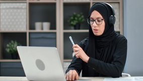 Modern Muslim business woman in headset talking online video call use laptop working studying remotely at home. Freelancer Asian female in black headscarf e learning internet education or helpline
