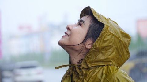 young asian woman wear yellow raincoat enjoy pouring rain standing on the road side, looking up to the sky while rain drops to her face smiling, rainy season weather climate, positive expression