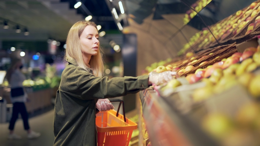 young blonde woman picks chooses fruits vegetables on the counter in supermarket. Female housewife shopping in market standing near vegetable department store with a basket in hands. Examines apple