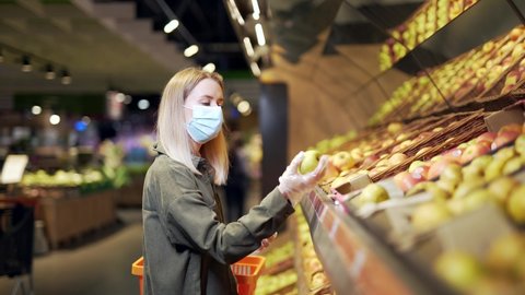 young blonde woman in a protective face medical mask picks chooses apple fruits vegetables on the counter in supermarket. Female shopping in market standing near department store with basket in hands