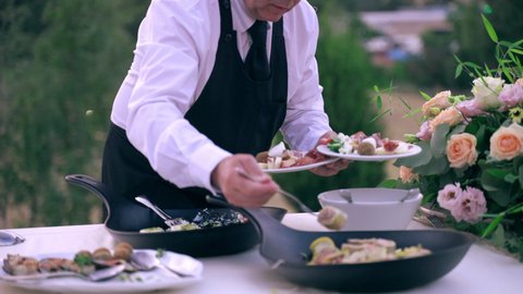 Professional waiter in uniform serving luxury wedding party outdoors at restaurant reception, servant putting variety of catering food on plates for guests. Banquet table full of snacks and tasty