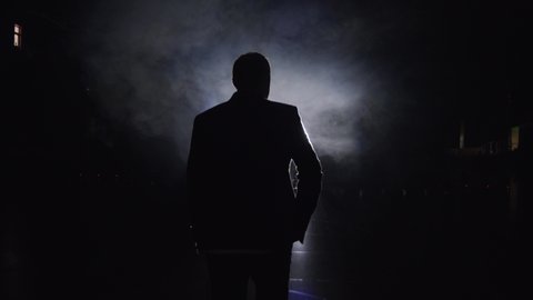 Silhouette of a man in a suit leaves the stage 4K. Mystical atmosphere, smoke, spotlight. Person in the dark