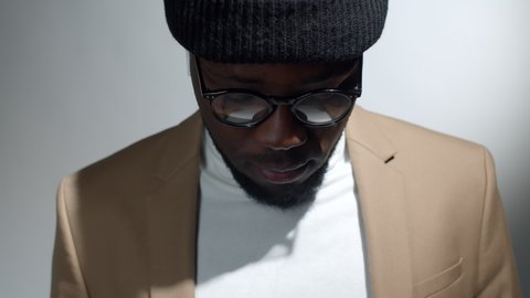 Tilt up portrait shot of young cheerful afro-american man in beanie hat, glasses, wireless earphones and smart casual outfit tilting up his head and smiling at camera while posing in studio