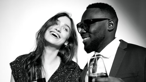 Black-and-white studio portrait of beautiful glamorous woman of Caucasian ethnicity and trendy Afro-American man holding wine glasses, smiling and posing together for camera