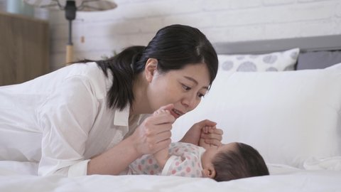 close up asian mother is leaning over and looking at her lovely young child with joy while holding its hands to touch her face on the bed at home.
