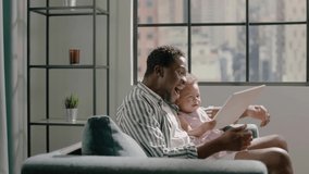 Afro-American father holds on knees toddler daughter playing with tablet in grey armchair with cushion against window in room slow motion - Powered by Shutterstock - Get 15% off with code: PIKWIZARD15