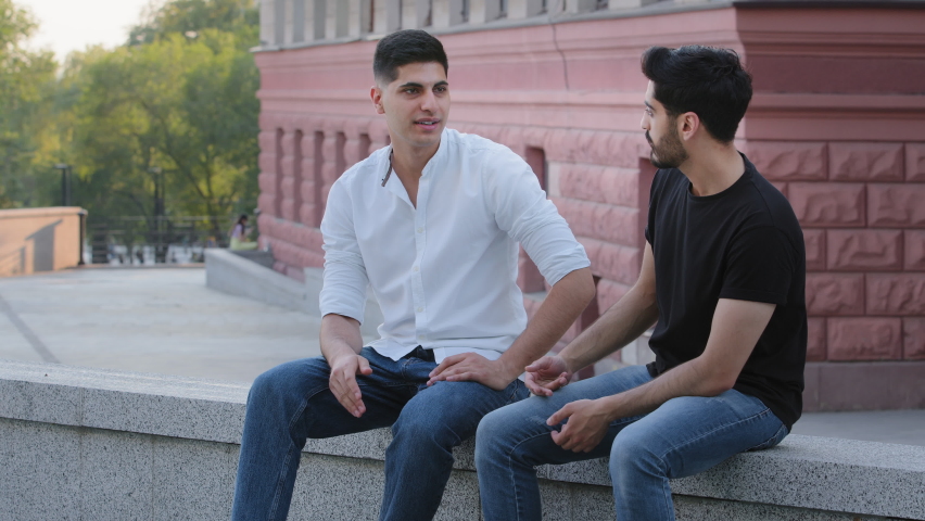 Two positive Indian Arab men sitting outdoors, having conversation, discussing educational issues or business problems. Carefree dark haired middle eastern boys friends sharing news enjoying weekend Royalty-Free Stock Footage #1077395336