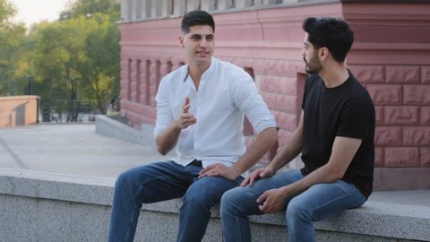 Two positive Indian Arab men sitting outdoors, having conversation, discussing educational issues or business problems. Carefree dark haired middle eastern boys friends sharing news enjoying weekend