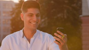 Smiling millennial Indian Arabic male student holding phone, making video call, talking to parents, girlfriend or wife, looking at camera outdoors. Young adult guy sends air kiss goodbye. Backlight - Powered by Shutterstock - Get 15% off with code: PIKWIZARD15
