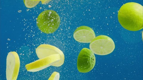 Close-up of falling ripe limes and lemons into sparkling water on blue background, making a cocktail of citrus fruits, drinking cold lemonade, shooting of carbonated water with floating sliced fruits.