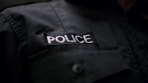 Closeup details of black police uniform. Policeman holding walkie talkie in pocket. Police badge attached to police jacket. Police lights of patrol car flashing on background 