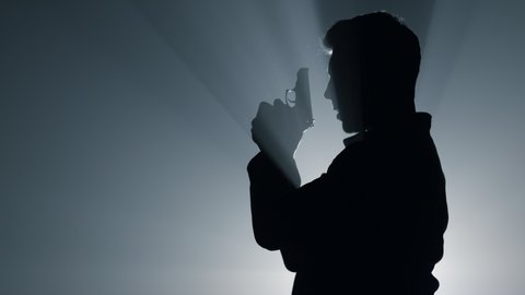 Silhouette of dangerous gunner shooting with pistol in floodlight background. Side view of focused man aiming with gun in darkness. Male criminal using firearm in dark. Crime concept.