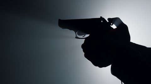 Silhouette of man hand holding gun indoors. Closeup unknown criminal arm cocking trigger in spotlight background. Unrecognizable male person shooting firearm in dark. 