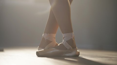 Close-up female feet in pointes standing up on tiptoes in backlit fog. Unrecognizable slim talented skilled Caucasian ballerina performing on stage in spotlight. Slow motion