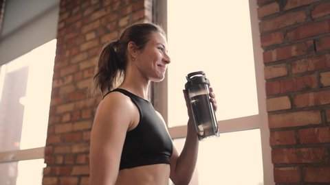Portrait of beautiful fit woman drinking water in a gym, after an intense workout, smiling. She is standing by a brick wall and looking out the window, smiling. Sport. Active lifestyle concept.