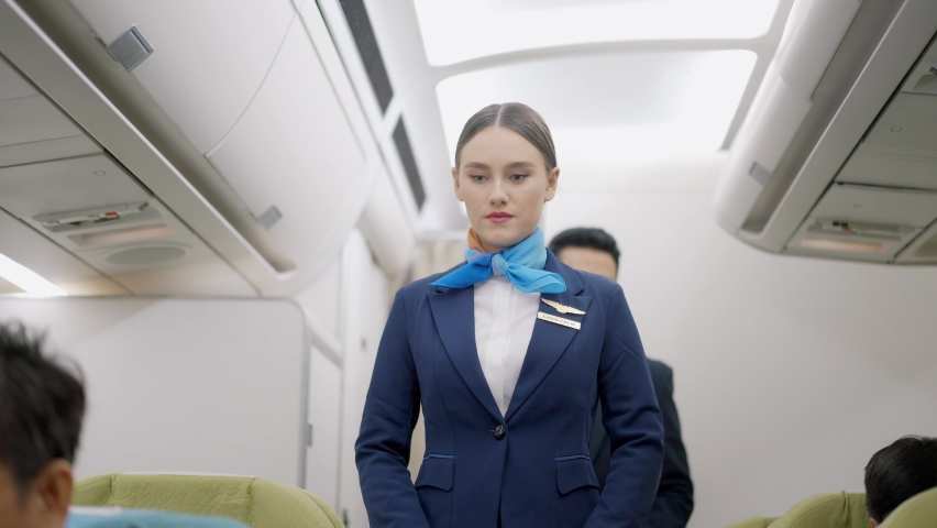 Caucasian female and Asian male flight attendants in uniforms walk along the aisle checking the passengers on safety standards with seat belt fastened, upright seat backs before airplane take off. Royalty-Free Stock Footage #1077425645